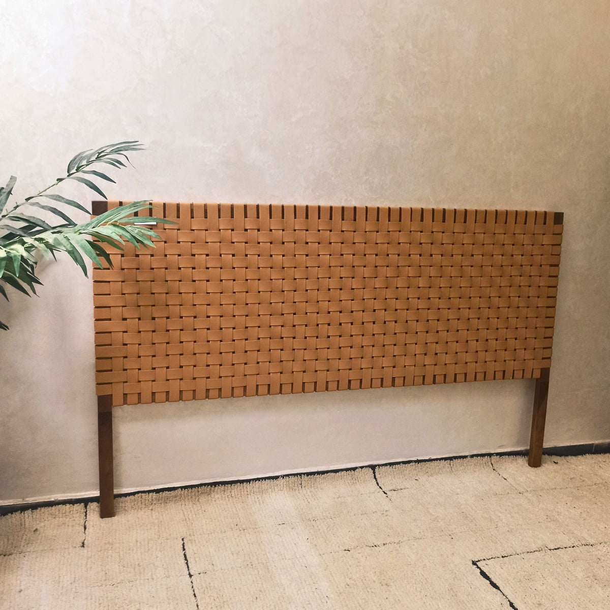 Woven walnut wooden headboard against a plain wall with a plant to the left.