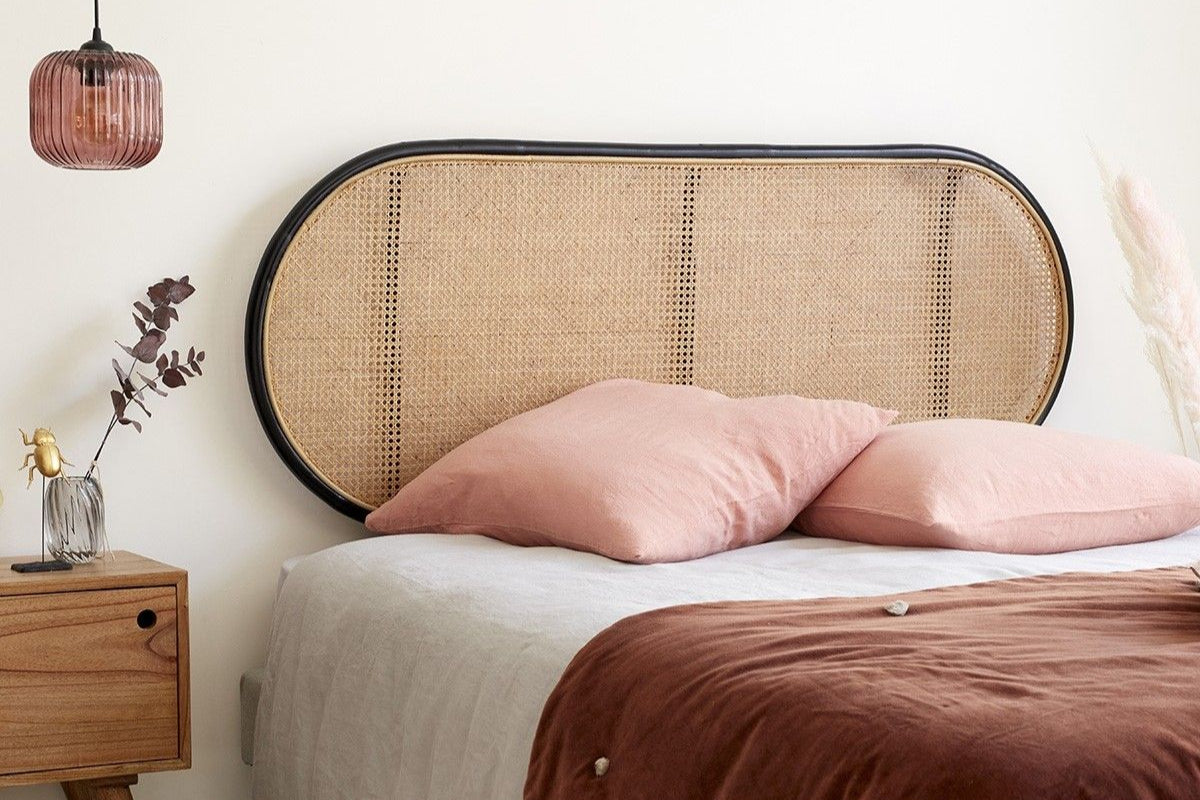 A rattan headboard with a wood frame, mounted on a wall above a bed with pink pillows and a brown duvet cover. The room has a cozy ambiance with a side table, lamp, and decorative items.